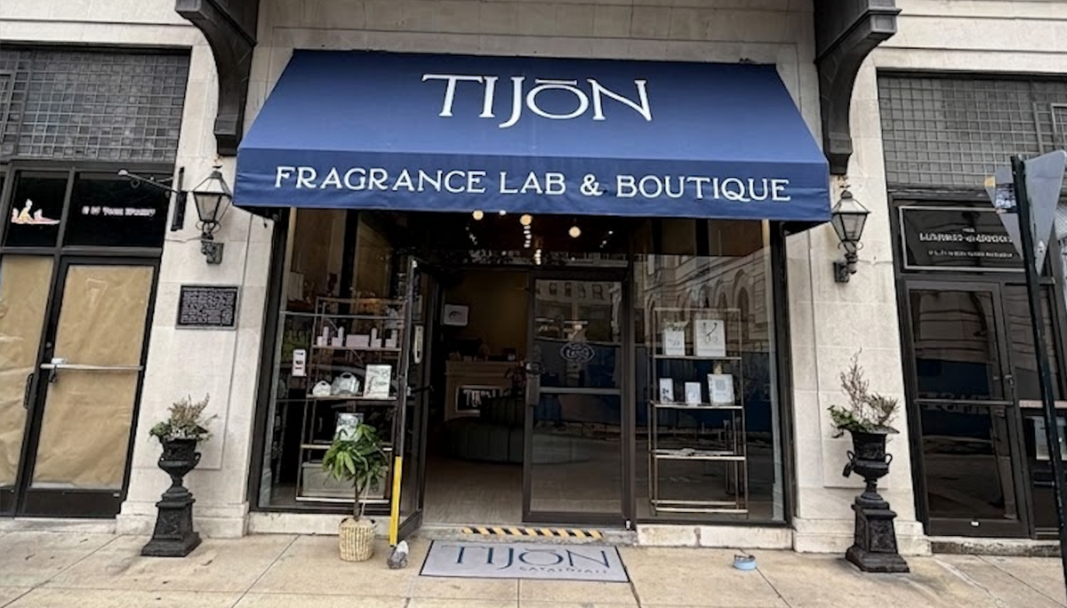 Front exterior of TIJON Fragrance Lab & Boutique, featuring a dark blue awning with white lettering, glass entrance doors, and stone facade walls.